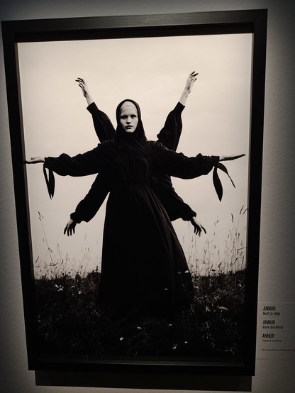 A woman is wearing a black dress and a scarf and it seems that she has six arms because people are standing behind her and only arms are visible. The black and white photo is a bit haunting.