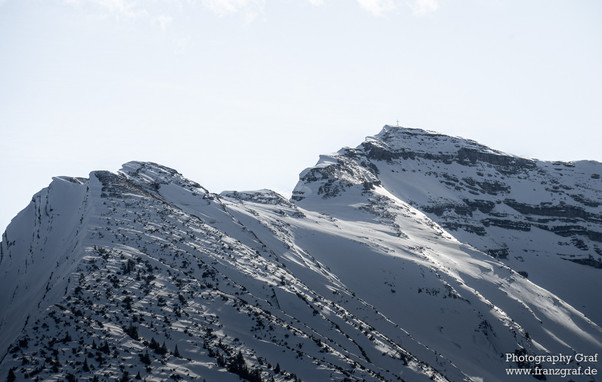 This image captures the raw beauty of nature in its most serene form. It features a majestic snow-covered mountain with a clear, radiant blue sky as the backdrop. The mountain's summit, slopes, and ridges are beautifully blanketed with a pristine layer of snow, making the glacial landform even more impressive. The terrain is also dotted with trees, adding a touch of life to the icy, white landscape. In the distance, one can see the outline of a mountain range, possibly the Alps, further enhanci…