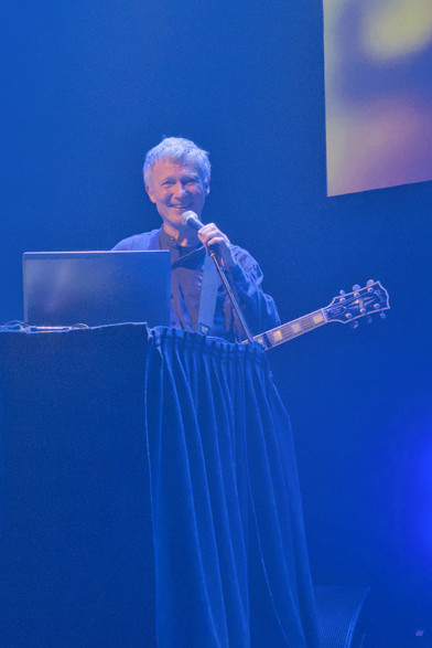 Michael Rother on stage in London's Barbican Hall. He has  a guitar slung around his neck and is stood behind a table. He is holding a microphone with one hand and smiling warmly at the audience.