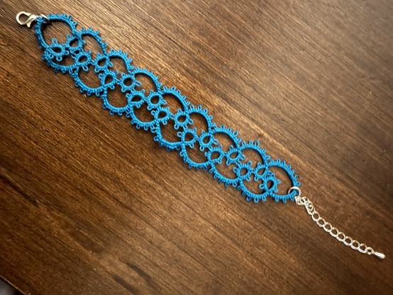 Blue tatted bracelet made of rings and chains with a silver clip and chain to close it, displayed on a wooden background. 