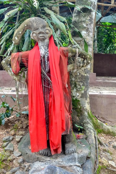 Photo of a statue of Ogun in the Sacred Grove Of Oshun in Osogbo, Nigeria. He is wearing a hat and bright red fabric is draped around his body.