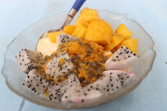 A glass breakfast bowl with a spoon with a blue handle in it, filled with fruit - white cubes of dragonfruit speckled with black seeds, deep orange-yellow cubes of mango, and passionfruit pulp in the middle.  The spoon has just a bit of white yoghurt from under the fruit on it.  