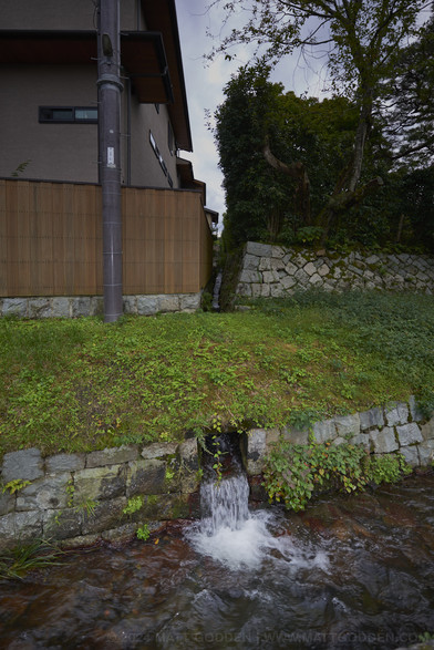 A stone-lined drain, flowing with a torrent of water, being met by a smaller, perpendicular stone-lined drain, which runs behind an apartment complex. The smaller drain runs under the nature strip - a grassy verge, before it empties into the main channel.