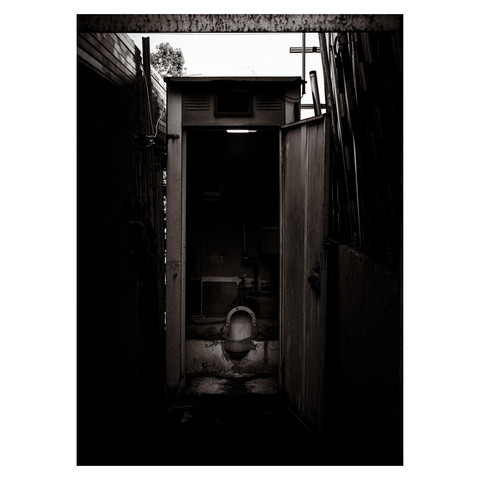 Presented here is a monochromatic glimpse into a tucked-away corner of daily life, where the simple yet vital facility of a traditional Japanese toilet stands. The open door invites the bold, while the darkness within promises a return to the basics of bygone eras, a whispered reminder of the functionality that underpins even the most modest of urban dwellings.