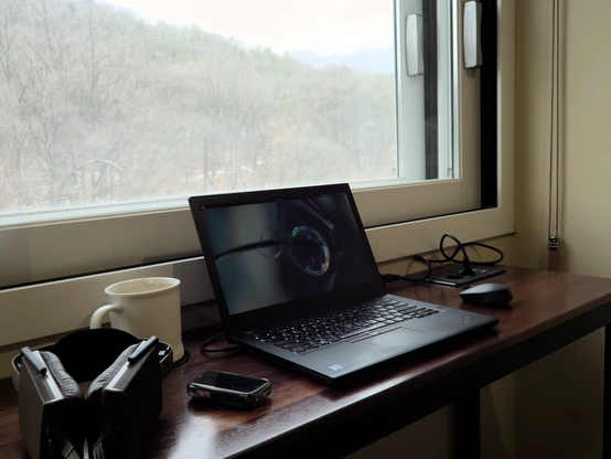 Photo of a Lenovo Thinkpad T480 sitting on a wooden counter top in front of a window. Through the windows you can see some remnants of snowy trees and mountains, though by this point in the afternoon much of the snow had already melted away. The Thinkpad's screen is on, showing an empty Fedora GNOME desktop as I flicked it over to an empty space as to not show my work contents.