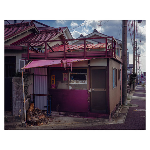 The image captures a quaint shop, now quiet and closed up, with remnants of its former life peering out from the window. The sun casts soft shadows across the structure, highlighting a few forgotten knick-knacks that linger as echoes of days filled with the bustle of customers and the ringing of the cash register. Now, it stands as a vignette of stillness, a pause in the urban narrative.