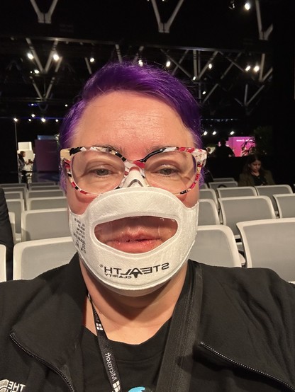 Woman with purple hair wearing a mask with a transparent inset that makes the mouth visible 