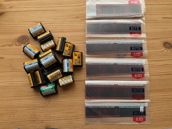 A handful of 35mm film cassettes to the left of several sheets of negatives in plastic sleeves, against an unfinished wood table top