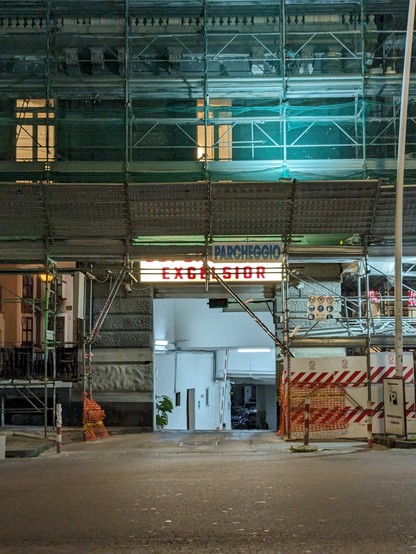 Shot of the neon-lit parking garage entrance of a hotel called Excelsior at night. The building is scaffolded and secured with green nets.
