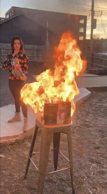 Valentina Gomez standing in what looks like a backyard firing a flame thrower directly at a bar stool, on top of which are placed a number of books which are now burning.