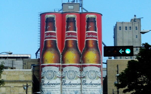 Photo of three silo-like buildings in Manitowoc painted with giant realistic-looking Budweiser beer bottles. In the foreground to the right is a traffic light with a left green arrow pointing at them.