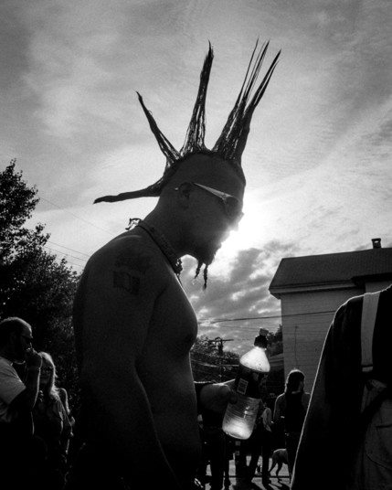 Silhouetted profile photo of a shirtless man with a spiked mohawk photographed from the waist up. He has two short braided strands of bear on his chin, is wearing sunglasses, and is holding a water bottle. There are other people and part of a house in the background.