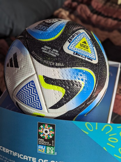 A white, blue, and shimmery black swirled Adidas Oceaunz soccer ball with neon yellow accents, nestled on a blue and white presentation box.