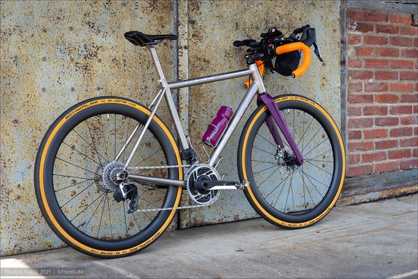 A road bicycle with a titanium frame and orange handlebars is leaning against a weathered wall with a rusted metal panel and brickwork.