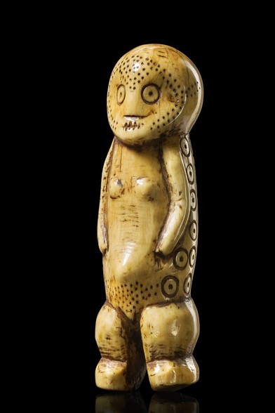 An ivory figurine of a standing woman with two strong legs, small breasts and slender arms. She is depicted with a prominent pubic triangle marked by dots and markings in the face at hat might be make-up or decorative scars.