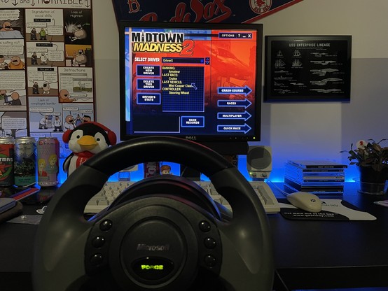 Computer monitor showing the main menu for the game Midtown Madness 2. In the foreground is a Microsoft Sidewinder Force Feedback Steering Wheel.