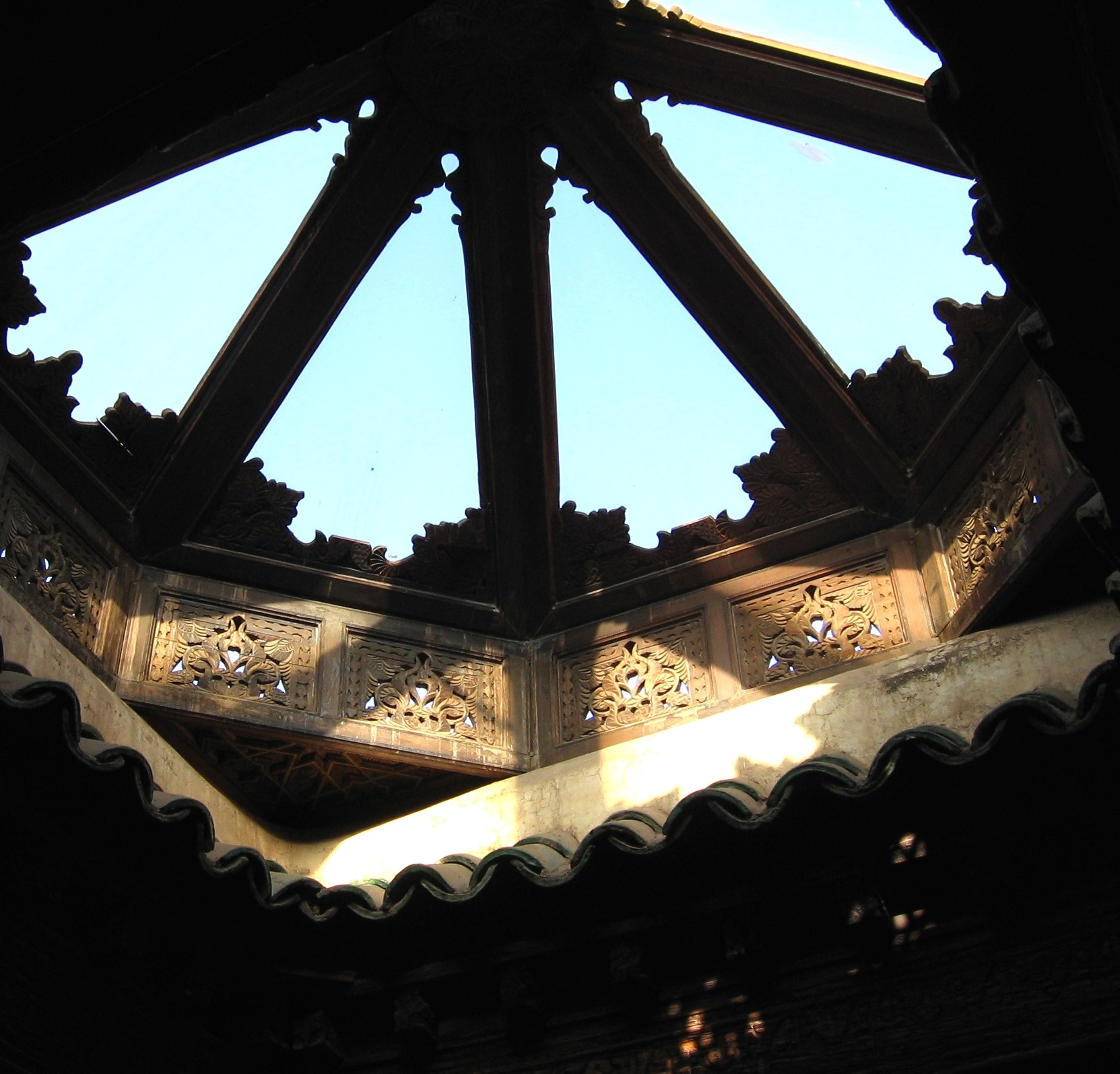 Photography of a very intricate window in a roof, circular with triangular sections through which blue sky is visible. The window frame is delicately engraved and has little pieces of mirror reflecting light.