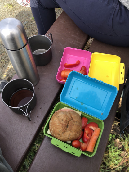 Versperzeit: Links eine Henkeltasse mit Kaffee, rechts daneben zwei bunte Vesperdosen, darin Paprikastreifen, Kirschtomaten, Möhren und ein belegtes Brötchen.