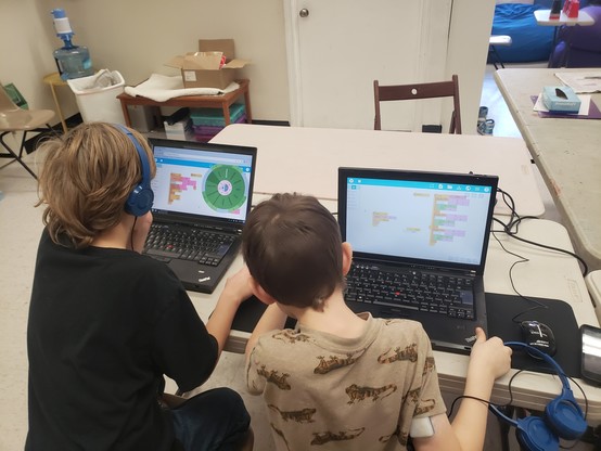 Two students at the computer, creating music with Music Blocks visual programming language. Both students have their backs to the camera. The student at left is wearing headphones and has a pie menu open. The student at right is looking at the other student's screen.