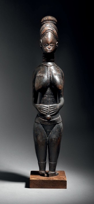 A large and elegant standing female statue made of wood with a black patina. She has her hands on her abdomen, full of roundness and delicacy in its smallest details. Her updo bun hairstyle is particularly fine and elaborate, a high forehead dominates a small and delicate face on a ringed neck. A long pearl necklace falls between the heavy breasts marked by horizontal scarification. The abdomen is barred by two long horizontal scarifications. A wide belt of beads rests on the loins and two ankl…