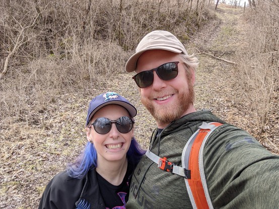 Selfie photo of Dan and I with the trail behind us. Both of us are wearing sunglasses and hats. I have facial piercings with blue and purple hair. Dan has strawberry blonde beard and hair. 