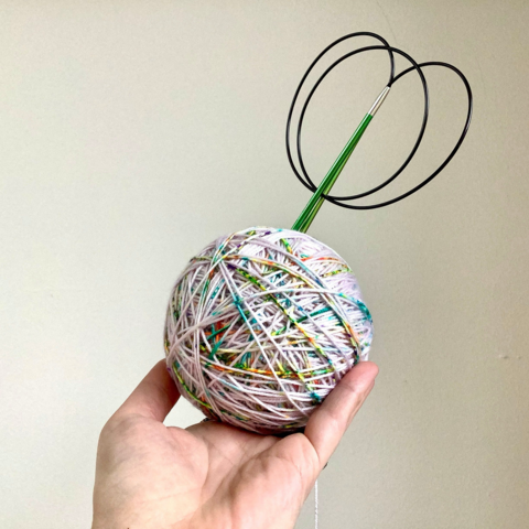A ball of pale pink yarn with multicolored speckles is held up against a white background. A green circular needle is inserted into the ball.
