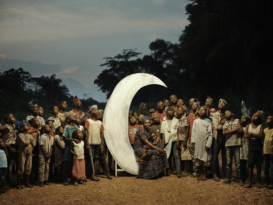 Photo of a woman sitting in a lunar crescent with a child, surrounded by other people. "The Woman on the Moon" from the series Congo Tales by Pieter Henket.