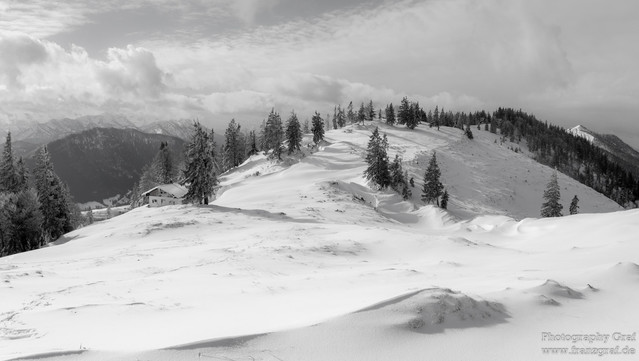 This image captures a serene snowy landscape, exuding the chilling beauty of winter. Dominated by a mesmerizing mountain range in the background, the scene is blanketed in a thick layer of snow. The sky is filled with a fluffy assortment of clouds, contributing to the overall white and grey color scheme of the image. A few trees, stark and bare, stand tall amidst this snowy terrain, adding a touch of contrast to the otherwise monochromatic view. Nestled somewhere within this wintry wonderland i…