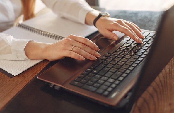 A woman typing at a keyboard. She is wearing a white blouse, with her arms resting on a blank notebook.