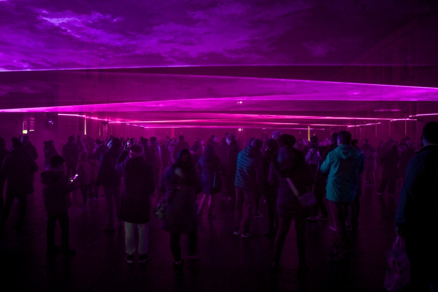 This image depicts a crowd of people gathered in an indoor space illuminated by vibrant purple and pink lights. The lights create horizontal lines across the image, giving the scene a dynamic and somewhat futuristic appearance. The people are dressed in casual attire, suggesting a public event or gathering.