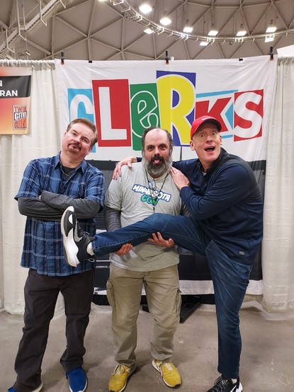 Me holding Jeff Anderson's right leg at Twin Cities Con 2023 while Bryan O'Halloran looks on disappointingly. I am wearing my Minnesota Con 2023 shirt from earlier in the year.