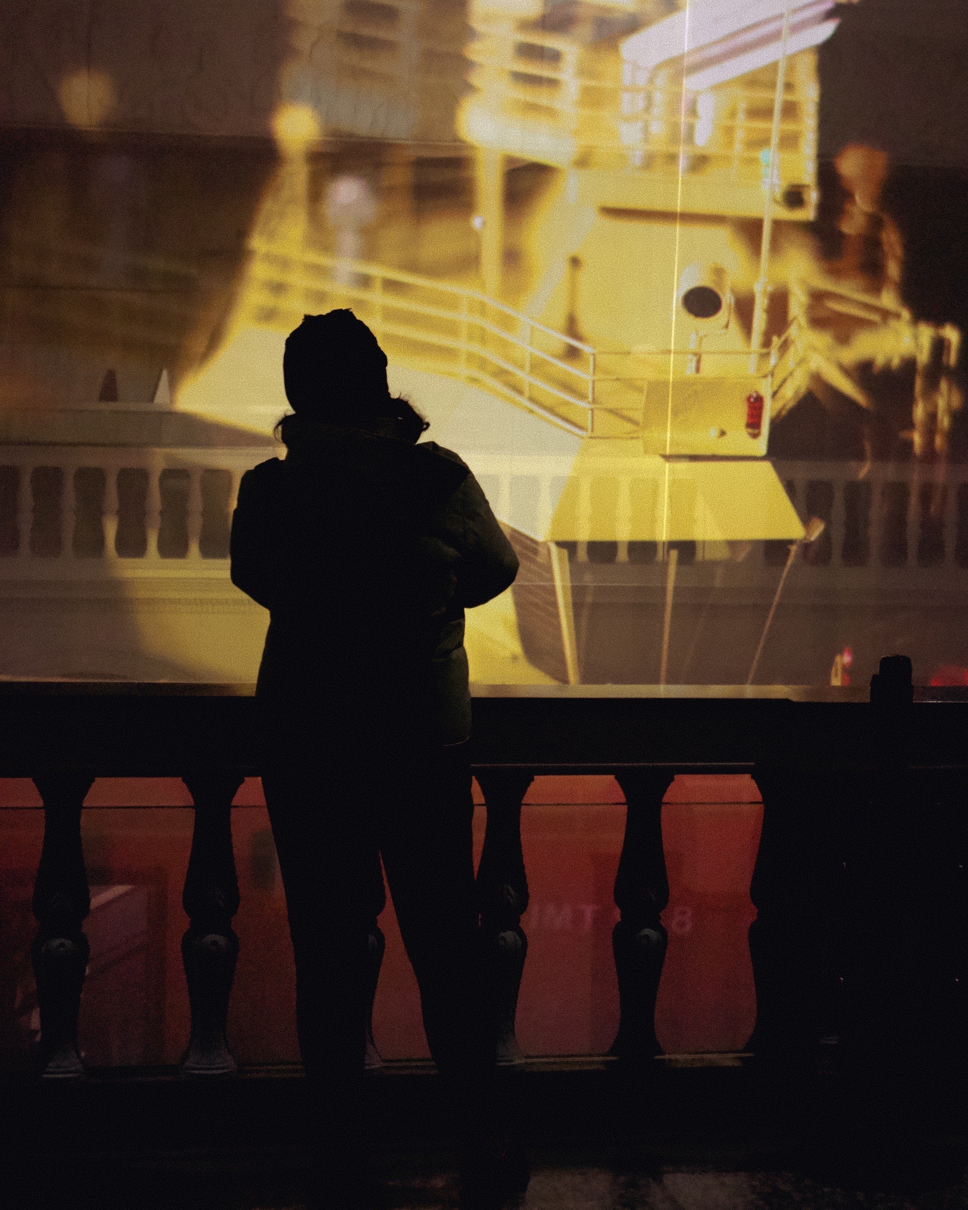 The image captures the silhouette of a person standing behind a balustrade, with a large, illuminated projection of a ship's structure cast upon them and the wall. The person appears to be photographing or observing the projected image, which includes details like stairs and railings on the ship. The reddish ambient lighting adds to the dramatic effect, creating a contrast between the silhouette and the golden hues of the projected ship.