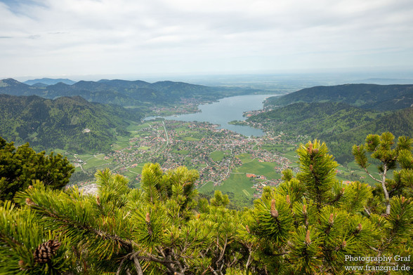 This photograph, taken by Franz Graf, features a stunning view of a lake and a town from a mountain. The landscape is dominated by shades of white, grey, and yellow, with a pop of vibrant green from the trees. The sky is a beautiful blue, dotted with fluffy clouds, and the overall scene is peaceful and serene. The mountain in the foreground adds depth and dimension to the image, while the lake and town in the background provide a sense of scale. The trees in the foreground, with their intricate…