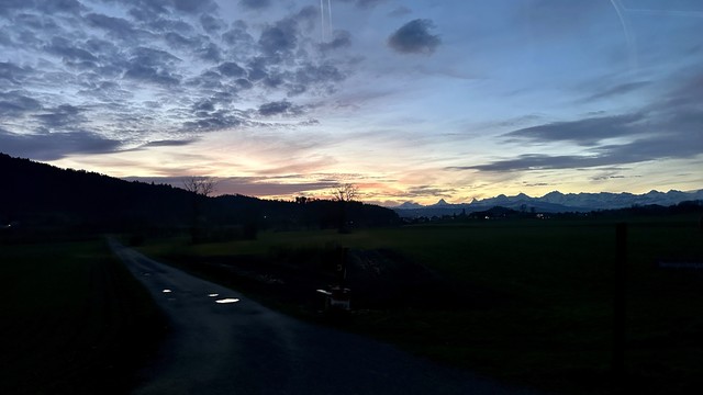 Unter leicht bewölktem Himmel sieht man in der Ferne die Silhouetten der Berner Alpen. Die Sonne schickt etwas gelb und Gold an den Horizont. Im Vordergrund glänzen ein paar Pfützen auf dem Kiesweg.