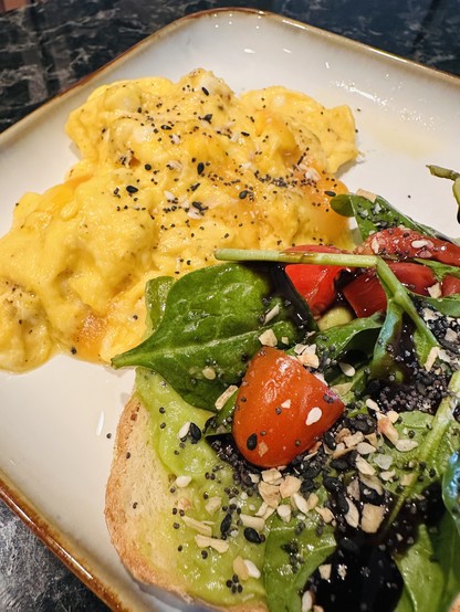 A small plate of softly scrambled eggs, top with everything bagel seasoning, and a slice of sourdough, avocado toast, top with spinach, tomatoes, and balsamic vinegar