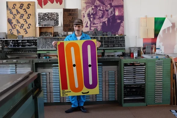 Swiss poster printer and typographer Dafi Kühne holding a poster with big number 100 on yellow background.