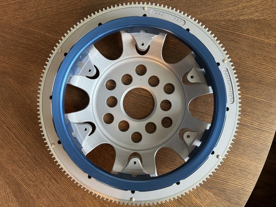 A grey flywheel with a gear around the edge of it. A blue pulley is attached to the front. 