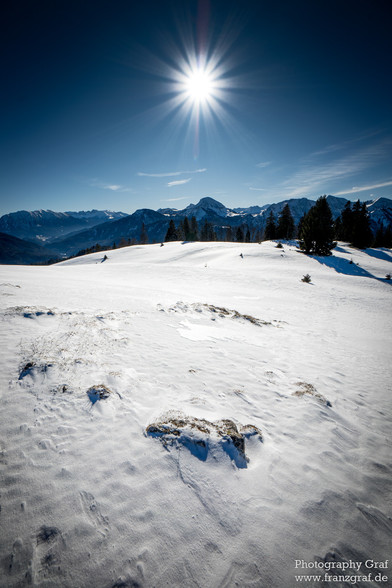 This image captures a breathtaking snowy landscape where the pristine white snow blankets the ground, creating a serene and tranquil scene. Tall trees, their branches laden with snow, stand majestically against the backdrop of majestic mountains that rise in the distance. The sky above is clear, suggesting a crisp winter day, with the sun possibly peeking through, casting a gentle glow over the entire scene. The landscape embodies the essence of winter, nature's freezing touch evident in the gl…
