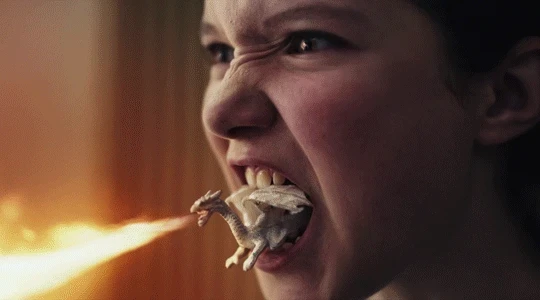 A scene from the film Blaze. A close-up of a girl's face, angry. In her mouth, between her teeth sits a small, white, fire-breathing ceramic dragon.