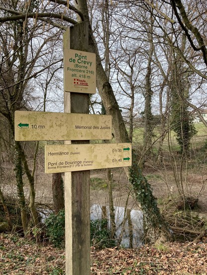 French walking signpost at the Pont de Crevy at border stone (“borne”) 216 in a strip of trees by the river. No leaves on the winter trees.