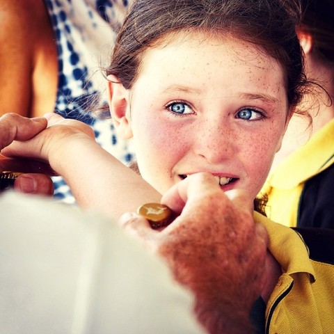 A close-up of a freckled young girl with blue eyes looking slightly away from the camera, her hand resting on an adult's hand in the foreground.