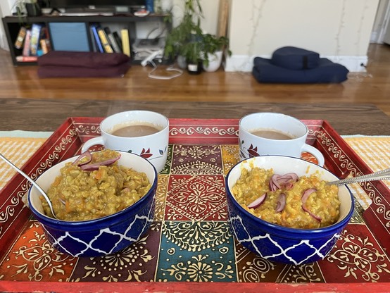 Two bowls of yellow colored oats garnished with red onions on a decorative tray accompanied by two cups of filter coffee, with meditation cushions and a bookshelf in the background.