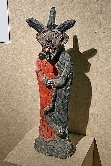 Painted concrete statue of Bosou with a pipe. He has three horns and a beard. In his hand he holds a snake. His robe is painted red, his principal colour.