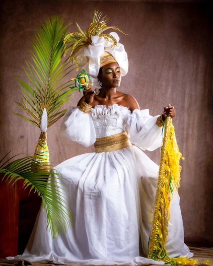 Photo of a black woman representing Ayizan Velekete. She is dressed in white and gold with a beautiful headdress an a glittering golden staff. Fringes of palm, her favourite plant, complete the composition.
