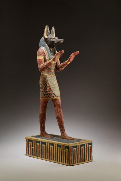 Statuette of jackal-headed Anubis made of plaster and painted wood. His head is black, his human body a bronze tan. He is wearing a wig, a beautiful kilt with a colourful pattern and golden arm rings. The work is dated to 332-30 BCE.