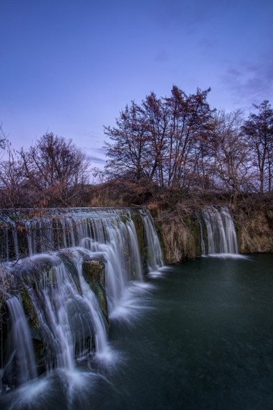 Se puede ver un azud o pequeña cascada con árboles y luz casi de la hora azul.

You can see a little waterfall with blue hour light.