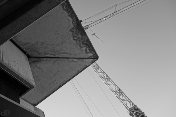 Schwarzweißfotografie
Diagonal von unten rechts ragt ein Kran in das Bild und nach oben rechts wieder heraus
Die Kabine und die Spitze des Krans ist von einer Ecke eines Betondachs verdeckt, an dem sich ein Muster aus Wasserflecken gebildet hat.
Das Dach ragt von der linken Seite in das Bild und verdeckt es bis zur Mitte