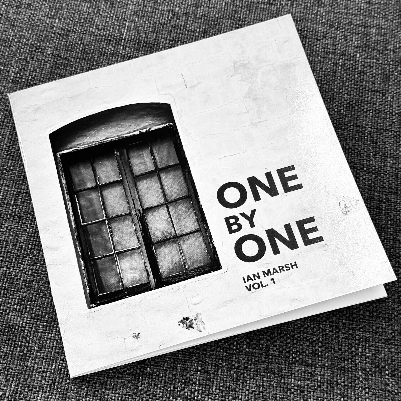 The image is a black and white photograph of a book with a textured cover. The book's cover features a realistic image of a window with multiple panes, some of which are slightly ajar, set into a white wall. Below the window, the book title "ONE BY ONE" is printed in large, bold, uppercase letters, followed by the name "IAN MARSH" in smaller, uppercase letters, and the text "VOL. 1" below the name. The book is lying on a gray fabric surface with a noticeable weave pattern, and there are small, white paint specks visible on the cover, particularly near the bottom edge, suggesting the book may be used or placed in a creative setting.
