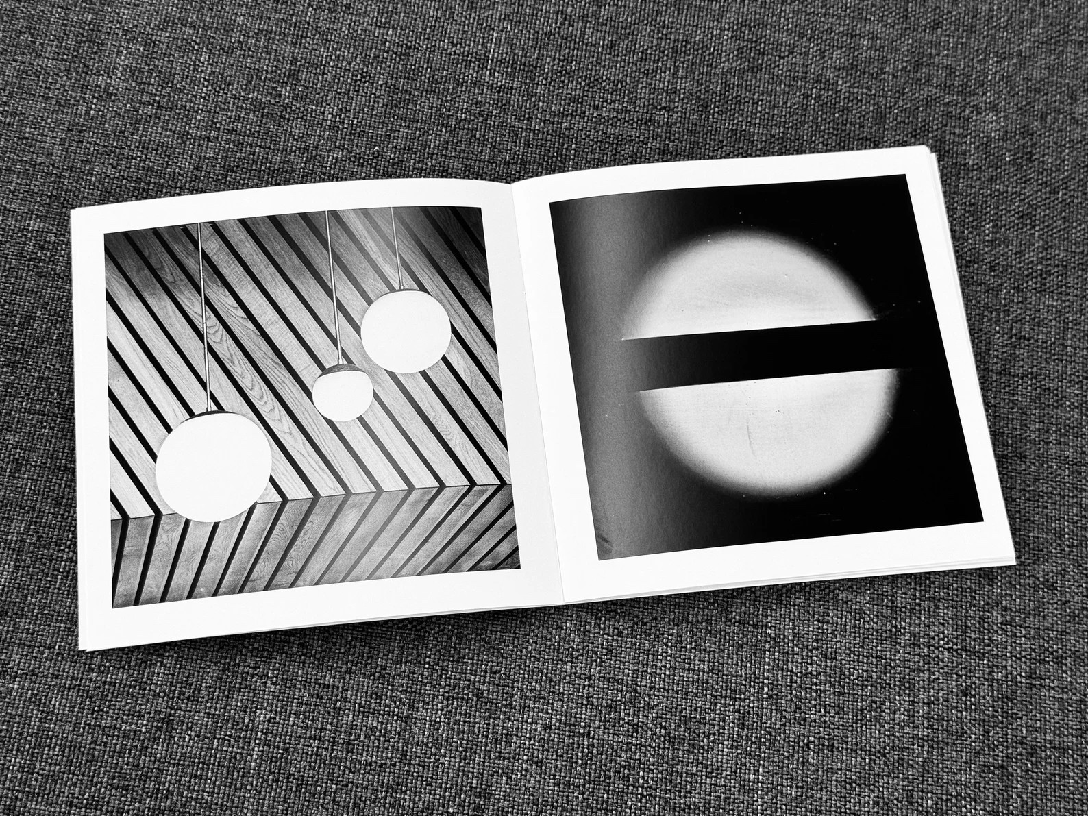
The image displays an open book on a gray fabric surface, showing two black and white photographs spread across both pages. On the left side, there is an abstract composition with three white circles, which appear to be suspended, against a background of dark, diagonally-striped patterns. On the right side, the photo depicts a large white circle with a dark horizontal line through its center, creating a bold contrast. The circle appears to be glowing, with a halo-like effect, against a dark background. Both images exhibit a play of geometry and contrast, focusing on shapes and the interplay of light and shadow.