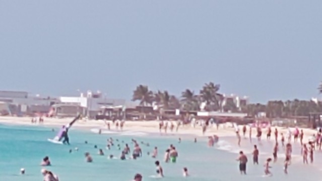 Geschwungene Küstenlinie, Telezoom Aufnahme mit Handy;
Viele Leute am und im Wasser, ein weißer Sandstrand, ein Windsurfer watet in tieferes Wasser 
Hintergrund mit relativ dezenten Gebäuden 